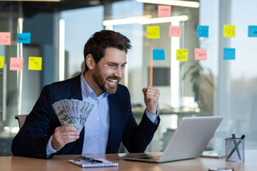 Successful businessman celebrating financial achievement at office desk holding cash and working on laptop. Smiling man expressing joy and excitement at work environment.