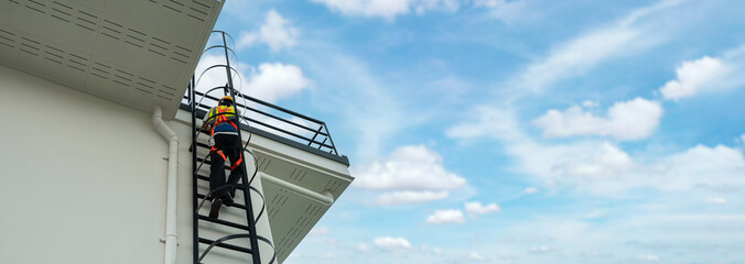 Industrial worker wearing safety harness climbing ladder to rooftop of building under clear sky, symbolizing occupational safety, construction industry, and workplace security standards. © AU USAnakul