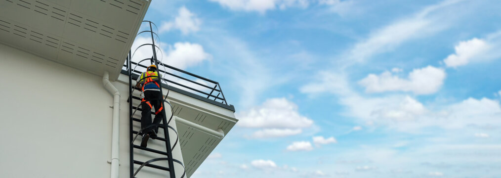 Industrial worker wearing safety harness climbing ladder to rooftop of building under clear sky, symbolizing occupational safety, construction industry, and workplace security standards.