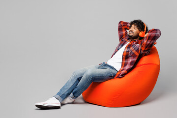 Full body young happy smiling Indian man he wearing red shirt casual clothes sit in bag chair listen to music in headphones isolated on plain grey color background studio portrait. Lifestyle concept.