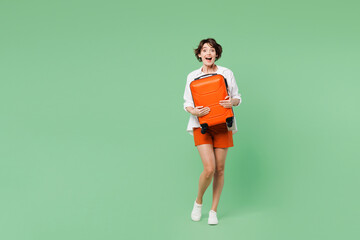 Full body happy surprised shocked traveler woman wear casual clothes hold suitcase bag look camera isolated on plain green background. Tourist travel abroad in free time rest. Air flight trip concept.