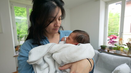 Mother cradling newborn baby, wrapped in white blanket, in modern living room, gazing lovingly at...
