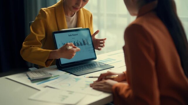 Two professionals analyze sales charts on a digital tablet, discussing performance metrics with documents on the table