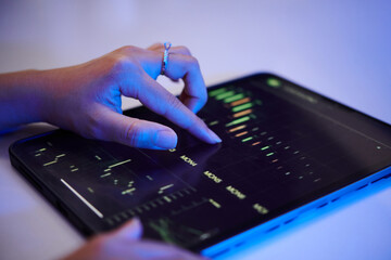 Hand using a tablet device for analyzing financial graphs and charts in a dimly lit setting, highlighting digital interaction and modern technology trends in finance