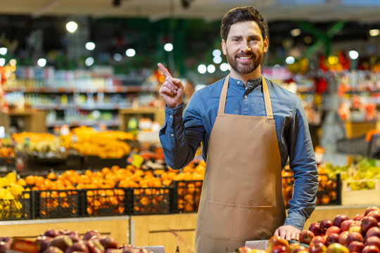 Grocery store employee in apron standing in produce section pointing finger. Bright fruits and vegetables on display creating colorful background. Image of customer service, retail, food, and market.