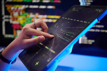 Close-up of person holding tablet displaying various stock market graphs and financial data with background of colorful code on screens creating dynamic atmosphere
