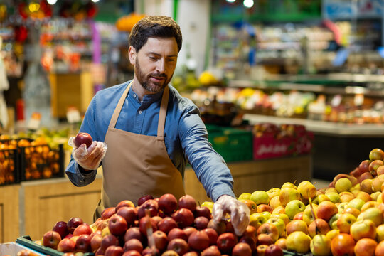 Male grocer wearing apron arranging apples in produce section of grocery store. Focus on fresh fruit display with vibrant colors. Concept of retail, grocery, and healthy food shopping. - Powered by Adobe