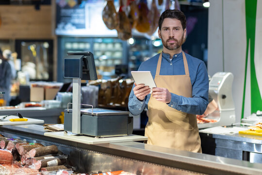 Male butcher in apron using tablet behind counter at deli shop. Display of various meats and sausages visible. Digital transformation in traditional retail setting