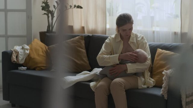 Medium shot of young Caucasian woman sitting on sofa holding baby during breastfeeding and gently stroking little head, feeling unconditional love from mother to child