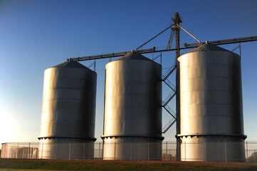 Three large metal grain silos stand in a row under clear blue skies at sunrise, with shadows and light highlighting their cylindrical shape in an agricultural storage facility