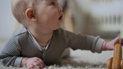 Close-up of adorable Caucasian baby wearing grey onesie nestling on carpet floor crawling to reach wooden car, curiously putting toy into mouth