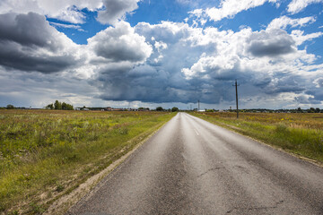 A long, winding road leads through idyllic fields, with towering clouds swirling above in a captivating display of nature's beauty on a warm summer afternoon.