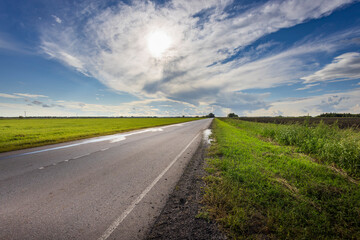 A tranquil road winds through vibrant green fields, illuminated by sunlight breaking through a stunning, dynamic cloudscape overhead.