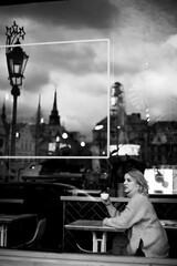 A woman sits at a table in a cafe, sipping coffee and looking out the window. The scene is set in a city with tall buildings and a street lamp in the background