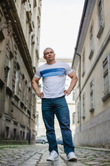 A man in a white shirt and blue jeans stands on a cobblestone street. He is looking to his right and he is in a good mood