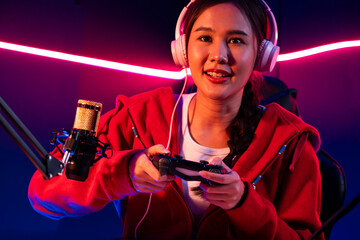 Host channel of beautiful Asian girl streamer with joystick playing online game wearing headphones pastel color talking with viewers media online. Esport skilled team players in neon room. Stratagem. © InfiniteFlow