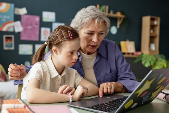 Senior woman guiding young girl with Down syndrome on laptop while sitting at a table in cozy home office, surrounded by educational materials - Powered by Adobe