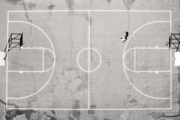 Direct overhead drone view of skateboarder on an empty basketball court on a sunny day, in black and white