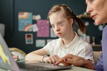 Girl with Down Syndrome engaging in home learning on laptop with adult supervision in background,...