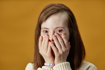 Portrait of young girl with Down Syndrome covering mouth with hands while smiling, captured against...
