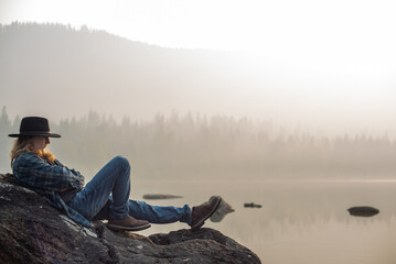 young man in flannel shirt and jeans, sitting by forest mountain lake