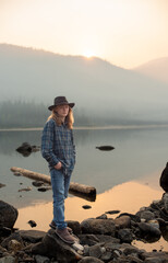 young man, wearing flannel shirt and jeans, standing by forest mountain lake