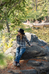 young man, wearing flannel shirt and jeans, sitting on rock by forest stream