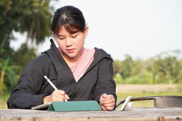 Young asian woman is seriously working remotely at outdoor area or park using a tablet. Natural...