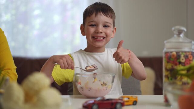 Close-up. A little boy looks at the camera and laughs, while holding a tablespoon with of breakfast cereal and yogurt. A child looks at the camera and smiles with a thumbs up, while sitting on a chair