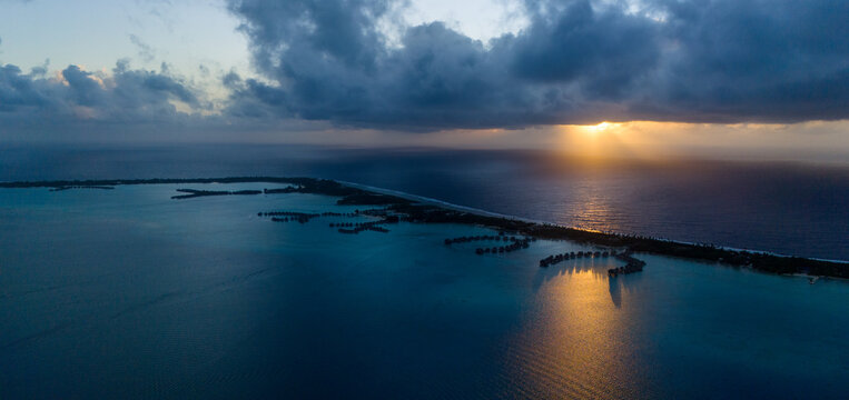 Early morning drone view of sunrise on the ocean horizon. Overwater bungalows can be seen on a motu near Bora Bora at tropical island resorts. Blue ocean water under dark clouds. 