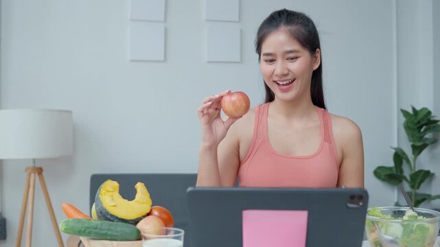 Woman smiles while enjoying an apple during an online meeting from home. Fresh vegetables surround her, blending healthy eating into her remote work lifestyle