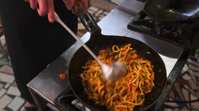 Appetizing preparation of lagman dish with white noodles, fried beef and vegetables seasoned with herbs close-up with stirring using a ladle in a wok over a gas stove.
