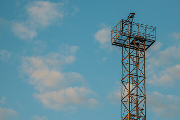 A tall metal tower with an observation platform stands against a clear blue sky dotted with soft clouds. The structure, made of metal beams, contrasts sharply with the tranquil sky