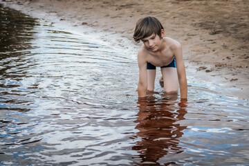 Fototapeta premium A young boy kneels in shallow water near the sandy shore, smiling softly as he engages in playful exploration. He wears blue swim trunks with colorful stripes, capturing the essence of a carefree