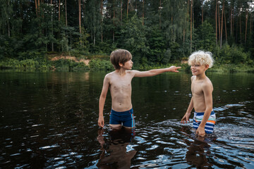 Fototapeta premium Two young boys stand in a calm river, surrounded by lush green forest. One boy points something out to the other, as they both enjoy the refreshing water. The image captures a moment of childhood