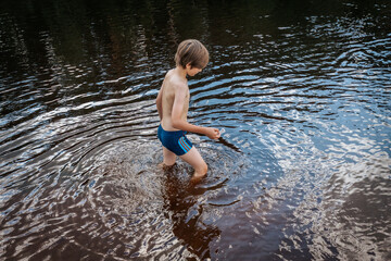 A young boy is seen crawling through the shallow water of a tranquil river, surrounded by lush greenery. His reflection is visible on the water's surface, adding a sense of adventure and exploration