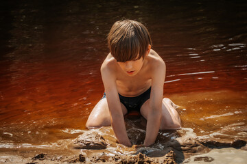 A young boy kneels in shallow water while holding a handful of wet sand. He is wearing blue swim trunks with orange stripes, and is focused on playing in the water. The image captures a moment of play