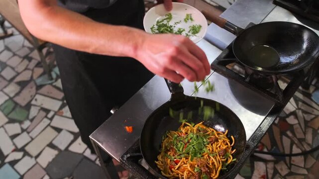 Male chef adds chopped greens to prepared uzbek dish lagman with white noodles and beef fried in wok on restaurant kitchen stove.