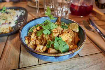 Pad Thai with chicken, peanuts, egg, lime sliced and green cilantro leaves in a blue bowl on a restaurant wooden table or at home
