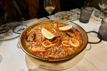Seafood paella with rice, prawns, red shrimp, mussels, squid rings, peppers, lemon wedges in its paella pan ready to serve on a white tablecloth on a restaurant table or at home