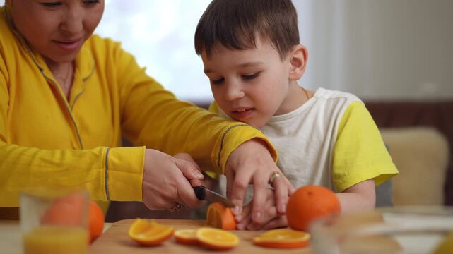 A mom and her little son together cutting an orange with a kitchen knife into slices, sitting at the table in the living room. A child's joy and laughter at the result of working jointly