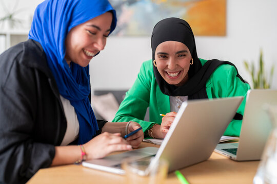 Two women in hijabs collaborate and smile while working on laptops at a desk in a modern office. The environment is bright and professional, reflecting teamwork and productivity