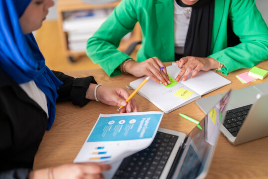 Two women in hijabs analyze data from charts and graphs while working at a desk in a modern office. Their focus and engagement highlight a professional and collaborative work environment.