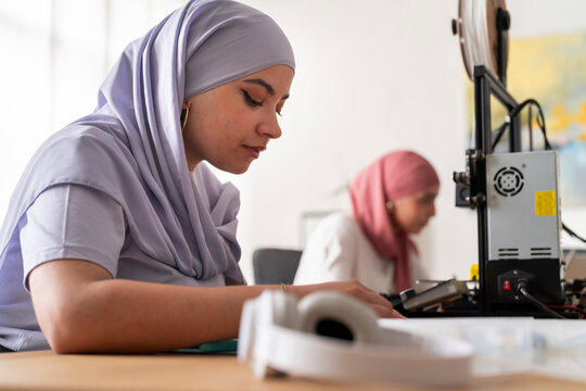 A woman in a light purple hijab is deeply focused on operating a 3D printer, adjusting its settings. The background shows another woman working, emphasizing a productive work environment.