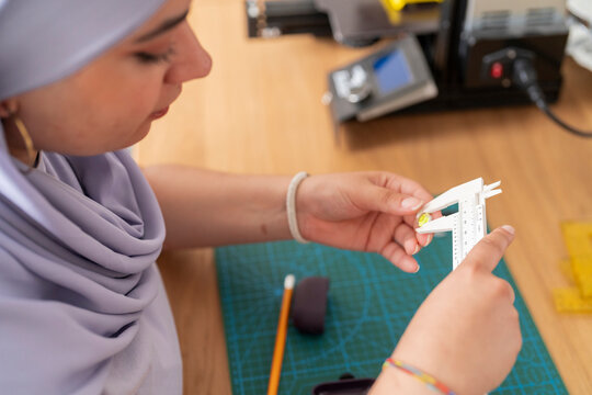 A woman in a light purple hijab uses a caliper to measure a small component for a 3D printing project. Her precise actions emphasize the importance of accuracy in engineering tasks.