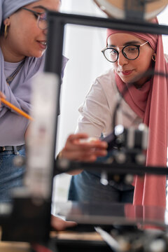 Two women in hijabs are seen adjusting the settings on a 3D printer, demonstrating teamwork and technical proficiency. Their focus on the machine underscores the complexity of the task.