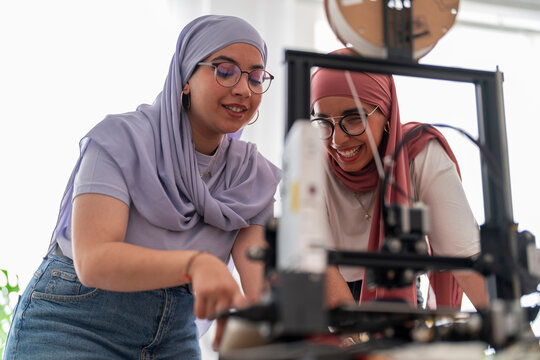Two women in hijabs collaborate on a 3D printing project, carefully examining the printer and its outputs. They are focused and engaged, showcasing teamwork and innovation in technology.