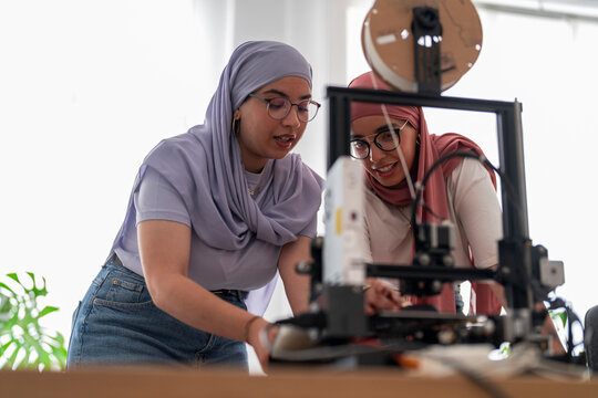 Two women in hijabs collaborate on a 3D printing project, carefully examining the printer and its outputs. They are focused and engaged, showcasing teamwork and innovation in technology.