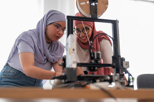 Two women in hijabs collaborate on a 3D printing project, carefully examining the printer and its outputs. They are focused and engaged, showcasing teamwork and innovation in technology.