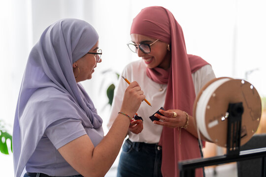 Two women in hijabs engage in a discussion about 3D printing components. They hold parts and tools, showcasing their involvement in the technical aspects of the project.